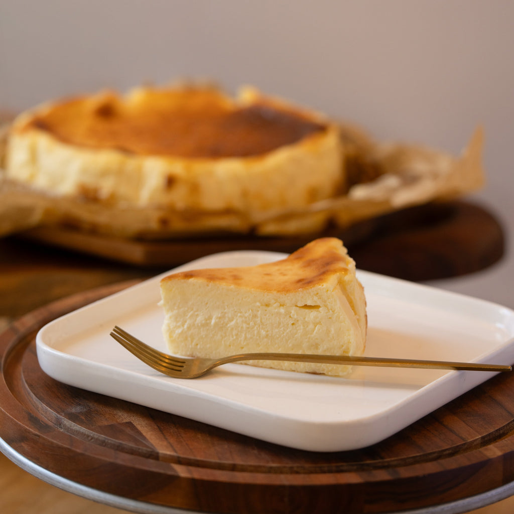 Slice of cheesecake on a white plate with a gold fork, whole cheesecake in the background.