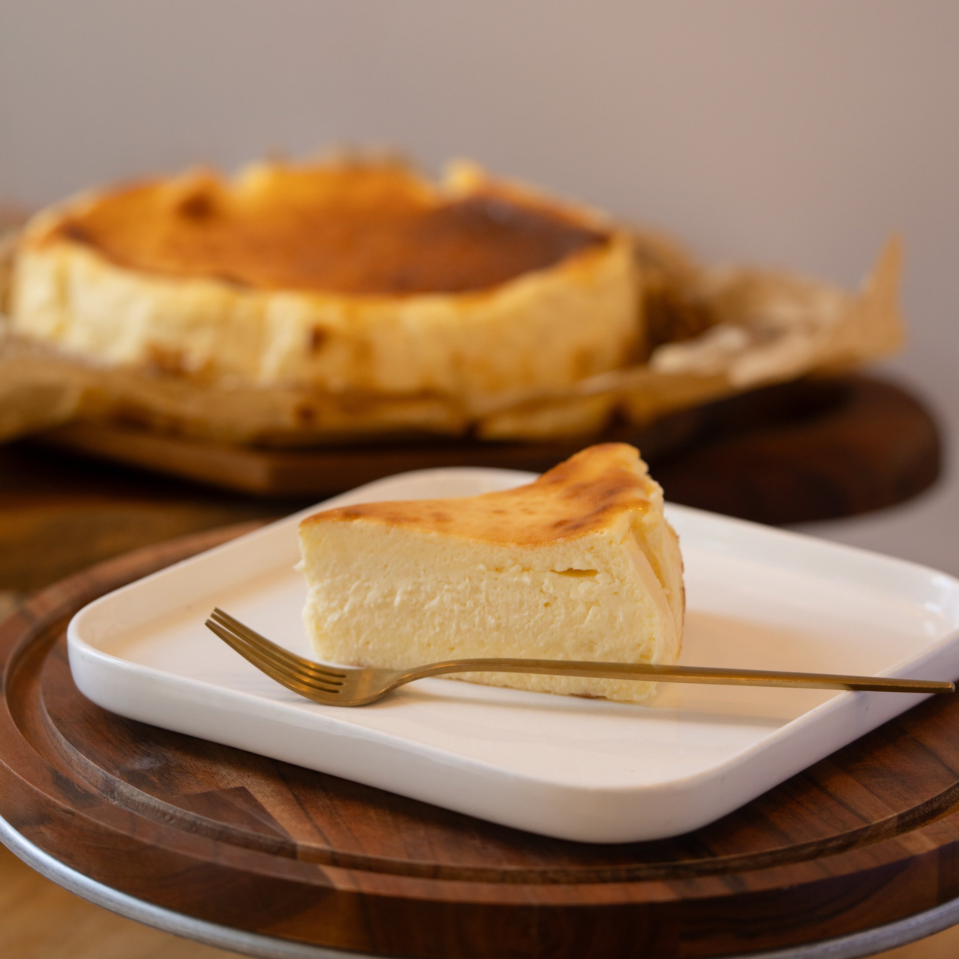 Slice of cheesecake on a white plate with a gold fork, whole cheesecake in the background.