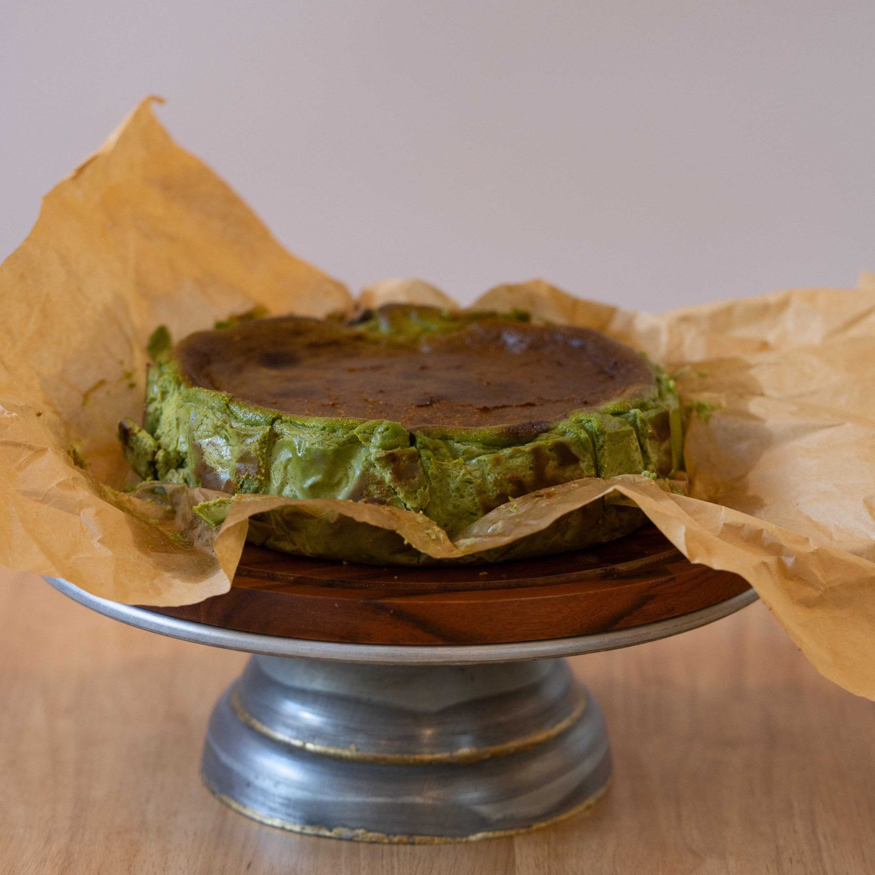 Green cake on a metal stand with brown paper underneath on a wooden surface.